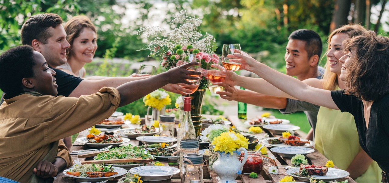 Six young people sit around a dining table outside and toast.