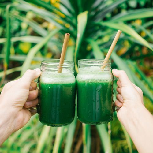 Two glasses with green liquid and bamboo straws are held up by two hands.