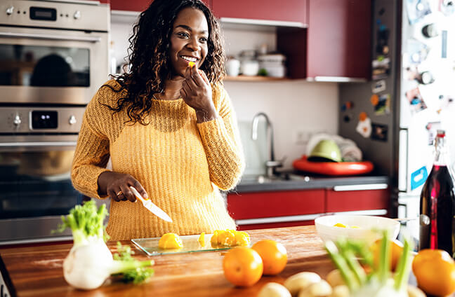 A young woman stands in the kitchen and tries a piece of an orange.