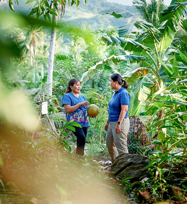 Zwei Frauen stehen in dicht bewachsenem tropischem Garten und sprechen über eine Kokosnuss vor grüner Hügelkulisse 