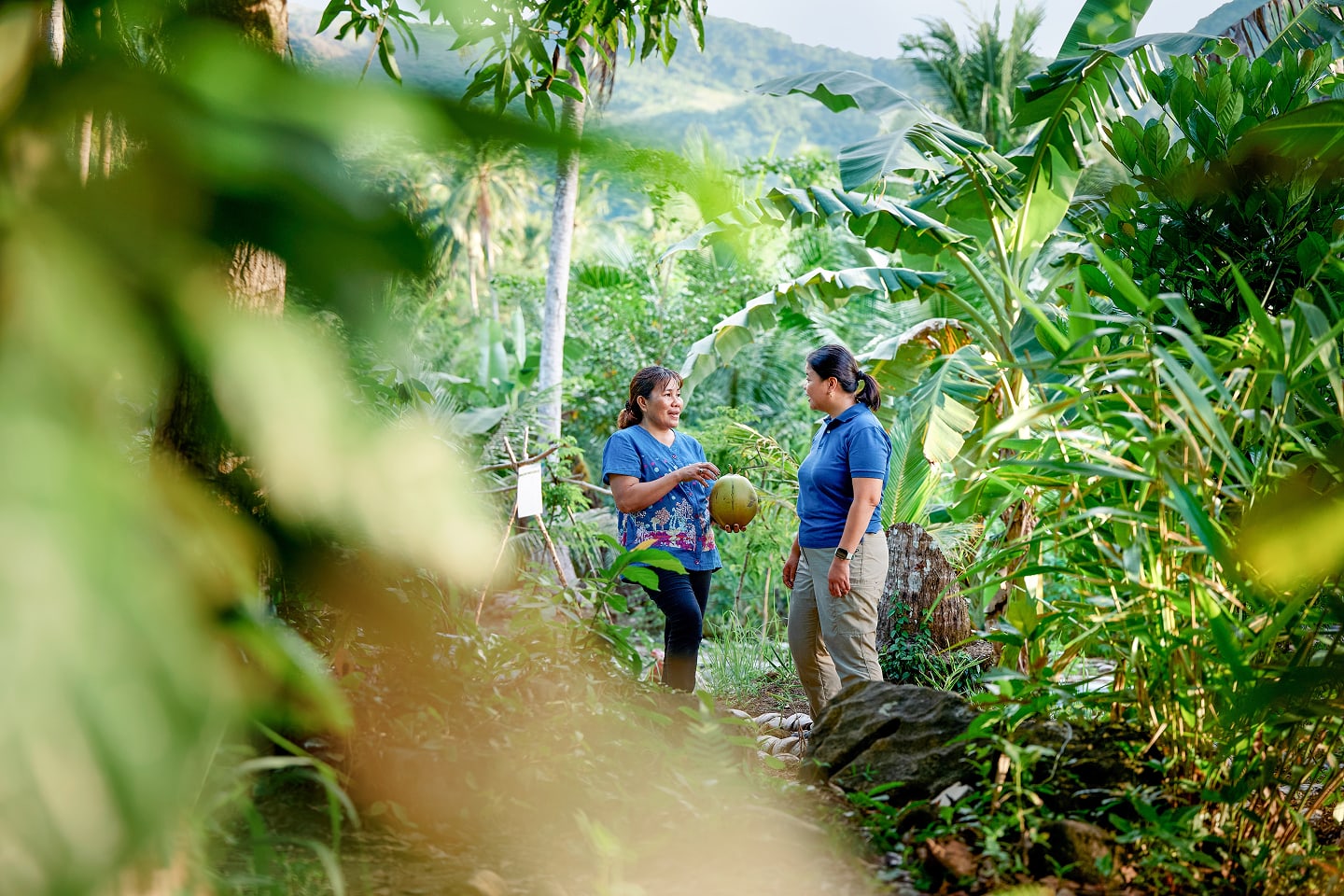 Zwei Frauen stehen in dicht bewachsenem tropischem Garten und sprechen über eine Kokosnuss vor grüner Hügelkulisse 