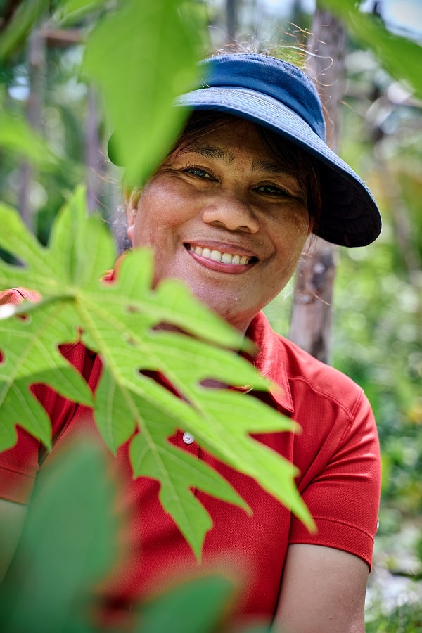 Frau mit blauem Visier lächelt zwischen grünen Blättern in tropischer Gartenumgebung und trägt ein rotes Shirt 