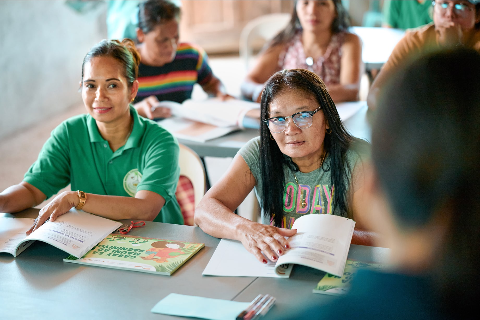 Gruppe Erwachsener sitzt an Tischen mit offenen Heften während einer Schulung und folgt der sprechenden Person vorn 