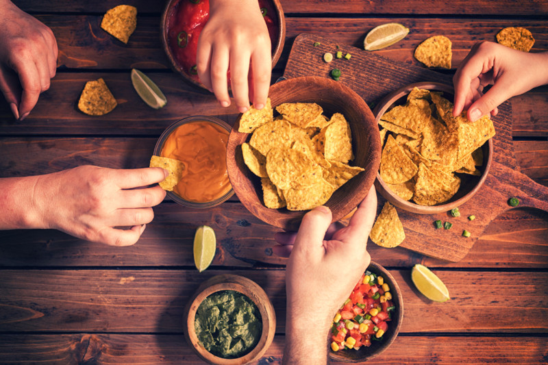 Mehrere Hände stecken Kartoffelchips in verschiedene Dips.